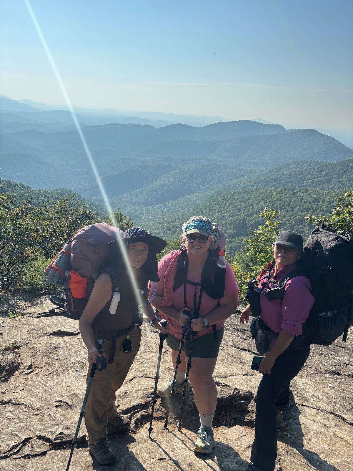 Three hikers with full packs at an overlook, mountains behind